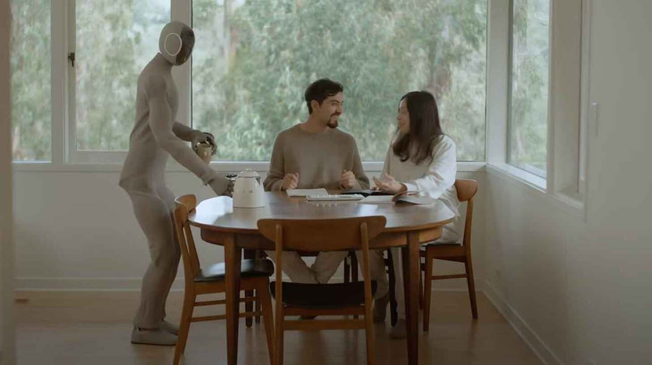 Paar inimest istub ümmarguse laua taga toas. Neid teenindab inimese moodi robot, kes hoiab kätel taldrikut. Aknast paistab loodusvaade.