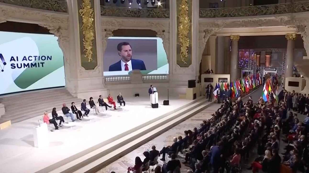 Luksuslikus ja avaras konverentsisaalis toimub 'AI Action Summit'. Laval istuvad mitmed inimesed, kes osalevad vestluses. Poodiumil seisab kõneleja ja tema pilt on suurel ekraanil taustal. Paremal pool on palju rahvuslippe rivistatud, publikus on hulgaliselt inimesi. Saali seinad on kaunistatud kuldse ornamenti ja keerukate kaunistustega.