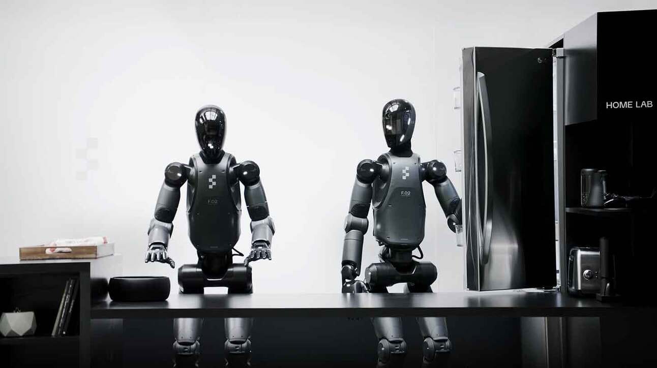 Kaks humanoidset robotit istuvad laua taga. Paremal olev robot avab külmkappi, mis on nende kõrval. Taustal on näha riiulit ja erinevaid esemeid.