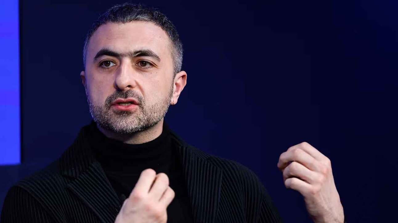Mees esineb sinise taustaga, käed on liigutuses. Tal on lühike habe ja tume juuksed, kannab musta kõrge kaelusega kampsunit.