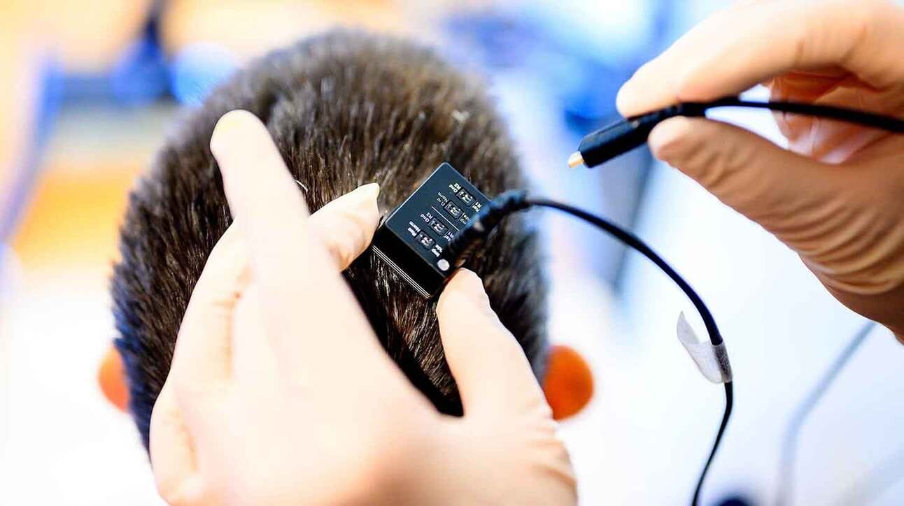 Käed kinnitavad väikese musta elektroonikaseadme inimese pea tagaküljele juustesse. Seadmele on ühendatud juhtmed.