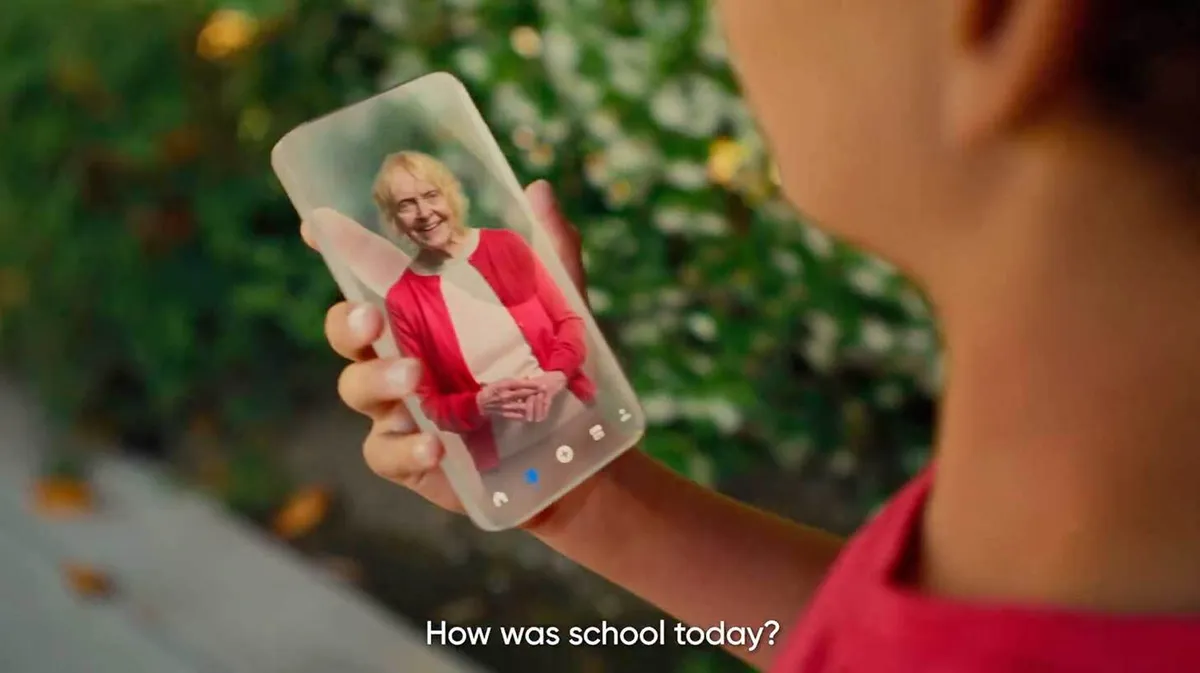 Laps hoiab käes nutitelefoni, millel kuvatakse videokõnes vanemat naist, kes naeratab ja kannab punast kampsunit ja valget särki. Tekst ekraani allosas: 'How was school today?'