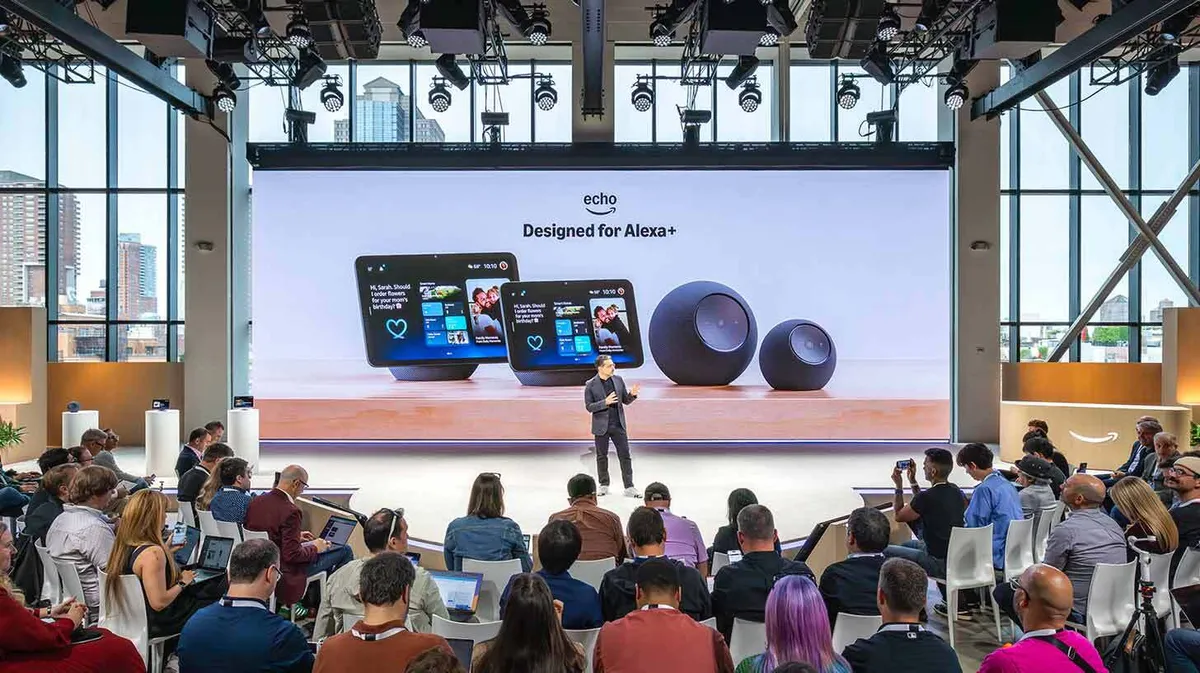 Konverentsisaal, kus esitletakse nutiseadmeid suurel ekraanil. Ekraanil on kaks ekraani ja kaks kõlarit, mille all on tekst 'Echo, Designed for Alexa+'. Publik istub ja kuulab esitlejat, kes seisab laval ekraani ees. Ruumi ümbritsevad suured aknad, kust paistab linnavaade.